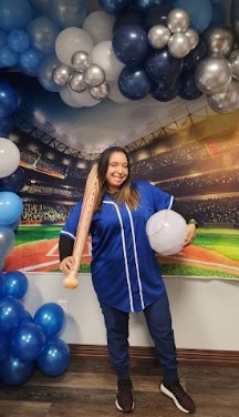 A full-body image of the prior employee standing in front of the stadium screen with blue balloons at her feet